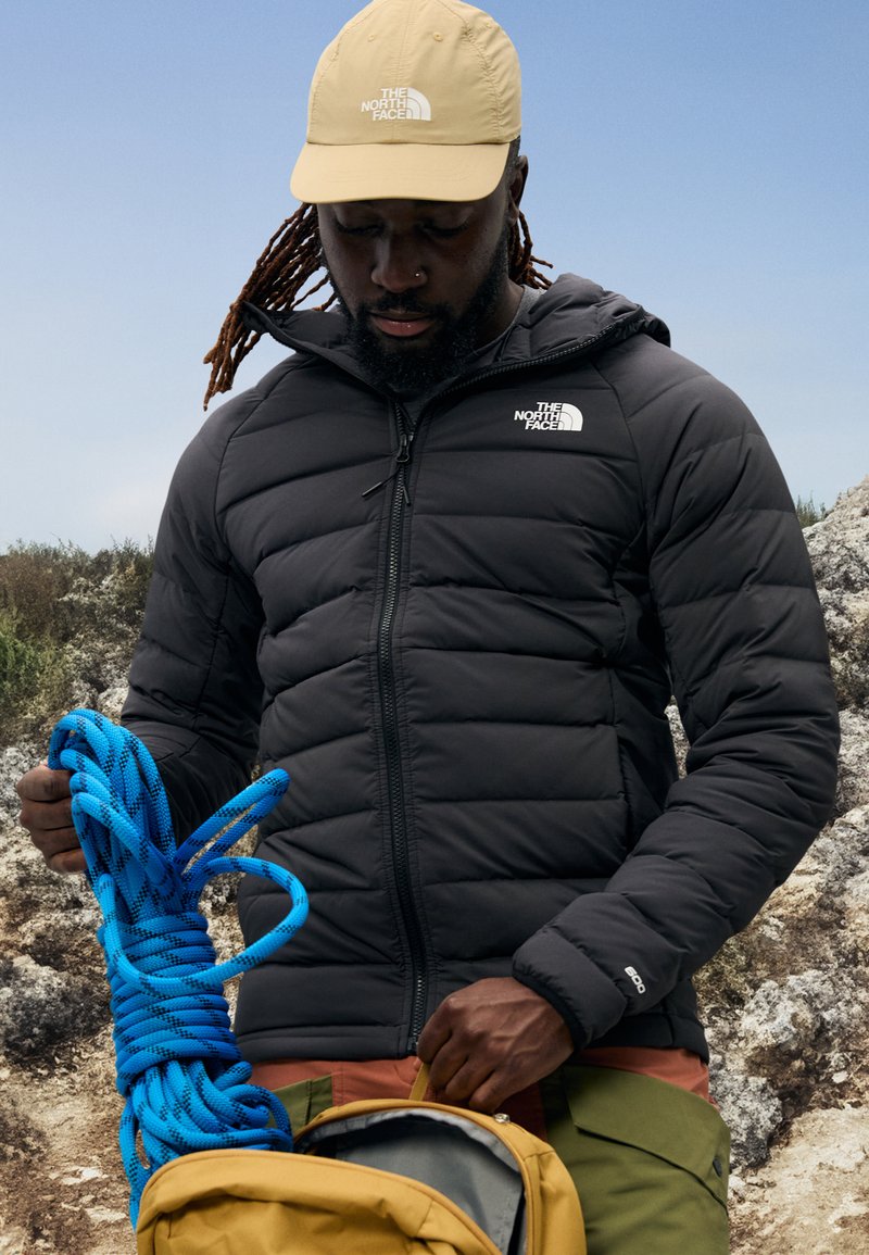 Giacca nera imbottita con zip e logo, cappellino beige, corda da arrampicata blu in mano, in piedi vicino a uno zaino marrone chiaro contro uno sfondo roccioso.