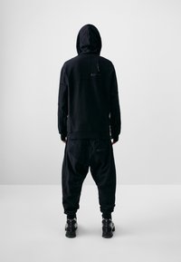 Schwarzer Kapuzenpullover mit strukturierten Ärmeln, kombiniert mit lockeren schwarzen Hosen mit Taschen und elastischen Bündchen. Schwarze Schuhe.