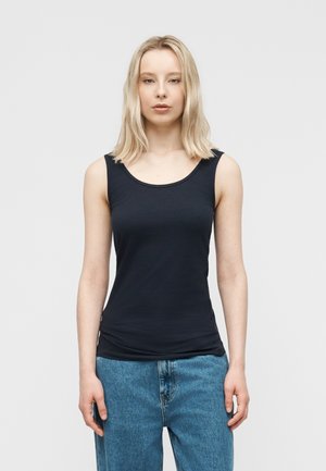 Blonde Frau trägt ein eng anliegendes schwarzes ärmelloses Oberteil und lockere blaue Jeans, steht vor einem schlichten hellen Hintergrund.