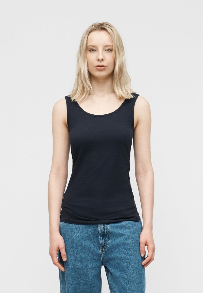 Ξανθιά γυναίκα που φοράει εφαρμοστό μαύρο αμάνικο τοπ και άνετο μπλε τζιν, στέκεται μπροστά σε απλό φωτεινό φόντο.