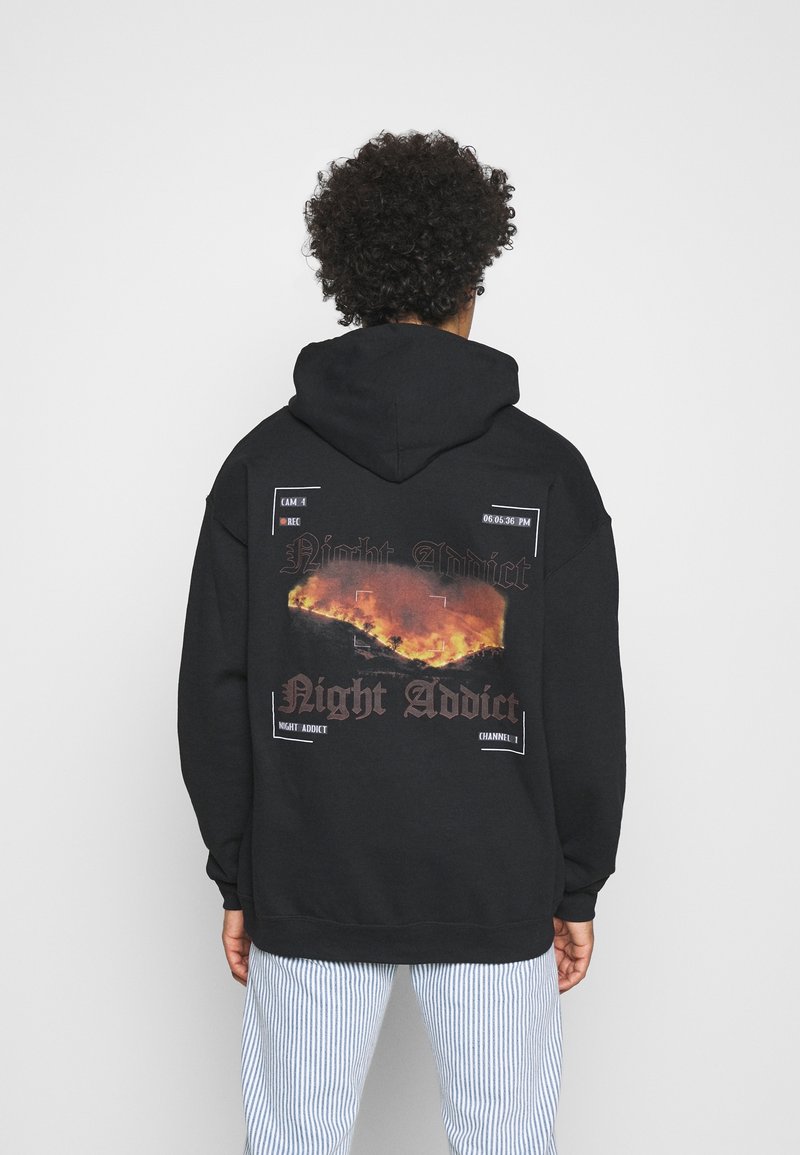 Persoon in een zwarte hoodie met een vlamgraphic en de tekst "Night Addict" op de achterkant, staande tegen een witte achtergrond.