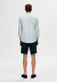 Camicia a maniche lunghe azzurro chiaro, pantaloni corti scuri, scarpe slip-on nere. La camicia ha polsini con bottoni e orlo curvo; il tessuto sembra liscio.