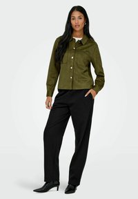 Olive green suede shirt jacket with two chest pockets, white buttons, paired with black tailored trousers and pointed black heels.