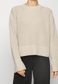 Maglione a maglia beige con motivo testurizzato, orlo ampio a costine e maniche lunghe leggermente svasate. Indossato con pantaloni scuri.