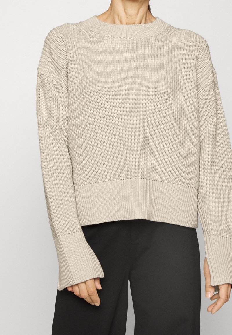 Maglione a maglia beige con motivo testurizzato, orlo ampio a costine e maniche lunghe leggermente svasate. Indossato con pantaloni scuri.