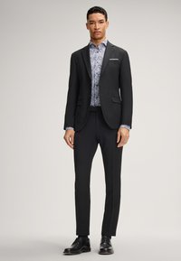 Completo nero con vestibilità affusolata, dotato di rever a punta e un fazzoletto bianco, indossato sopra una camicia blu a fantasia. Include scarpe eleganti nere.