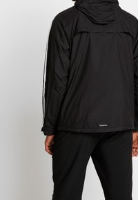 Svart vattentät jacka med huva, som har vita sidostripor, elastiska ärmslut och en synlig logotyp i nederkant på ryggen. Slät textur.