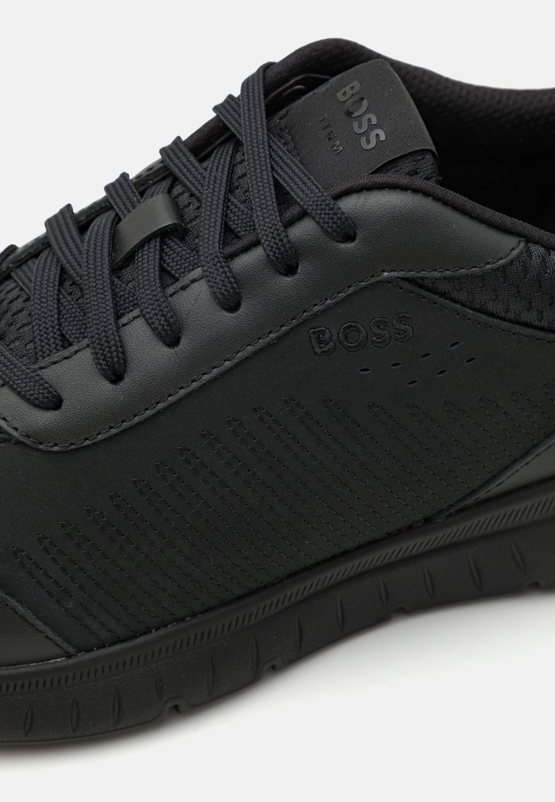 Zapatilla deportiva negra con parte superior sintética, panel lateral texturizado, cuello acolchado y marca en tono. Presenta cordones planos y una suela flexible.