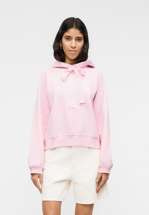 Femeie purtând un hanorac roz deschis cu un arc mare pe decolteu și pantaloni scurți albi, stând în fața unui fundal simplu.