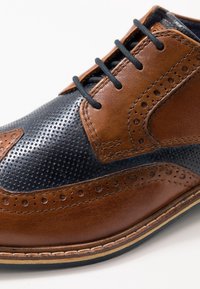 Chaussure brogue à lacets avec un design bicolore en cuir perforé bleu foncé et cuir lisse marron, ornée de broderies décoratives et de perforations.