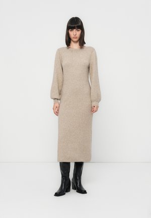 Robe en tricot beige à manches longues avec un col rond et une silhouette ajustée, mettant en valeur des manches bouffantes et une longueur mi-mollet. Bottes noires à talons.