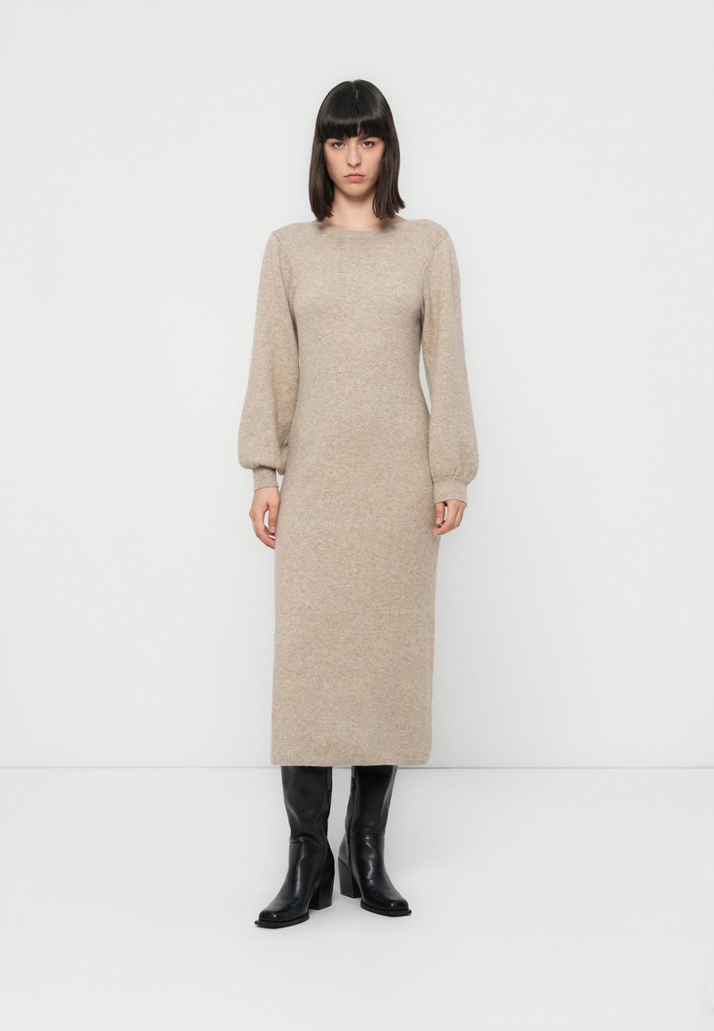 Robe en tricot beige à manches longues avec un col rond et une silhouette ajustée, mettant en valeur des manches bouffantes et une longueur mi-mollet. Bottes noires à talons.