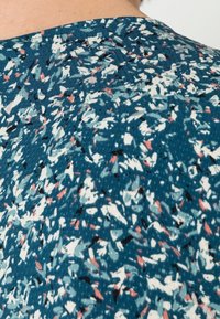 Kortärmad topp i teal tyg med ett texturerat, abstrakt mönster som har vita, rosa och svarta accenter. Rundad halsringning, avslappnad passform.