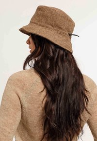 Cappello a secchiello marrone a spina di pesce realizzato in tessuto testurizzato, con una ampia visiera e un piccolo dettaglio decorativo con una fibbia nera di lato.