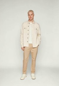 Μπεζ σακάκι με τσέπες μπροστά, φορεμένο πάνω από ένα λευκό πουκάμισο και μπεζ παντελόνι. Το άτομο στέκεται με λευκά αθλητικά παπούτσια σε ουδέτερο φόντο.