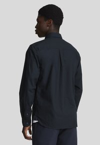 Camisa de manga larga negra hecha de algodón con cuello, costuras visibles y espalda ventilada. Presenta puños abotonados y un ajuste relajado.