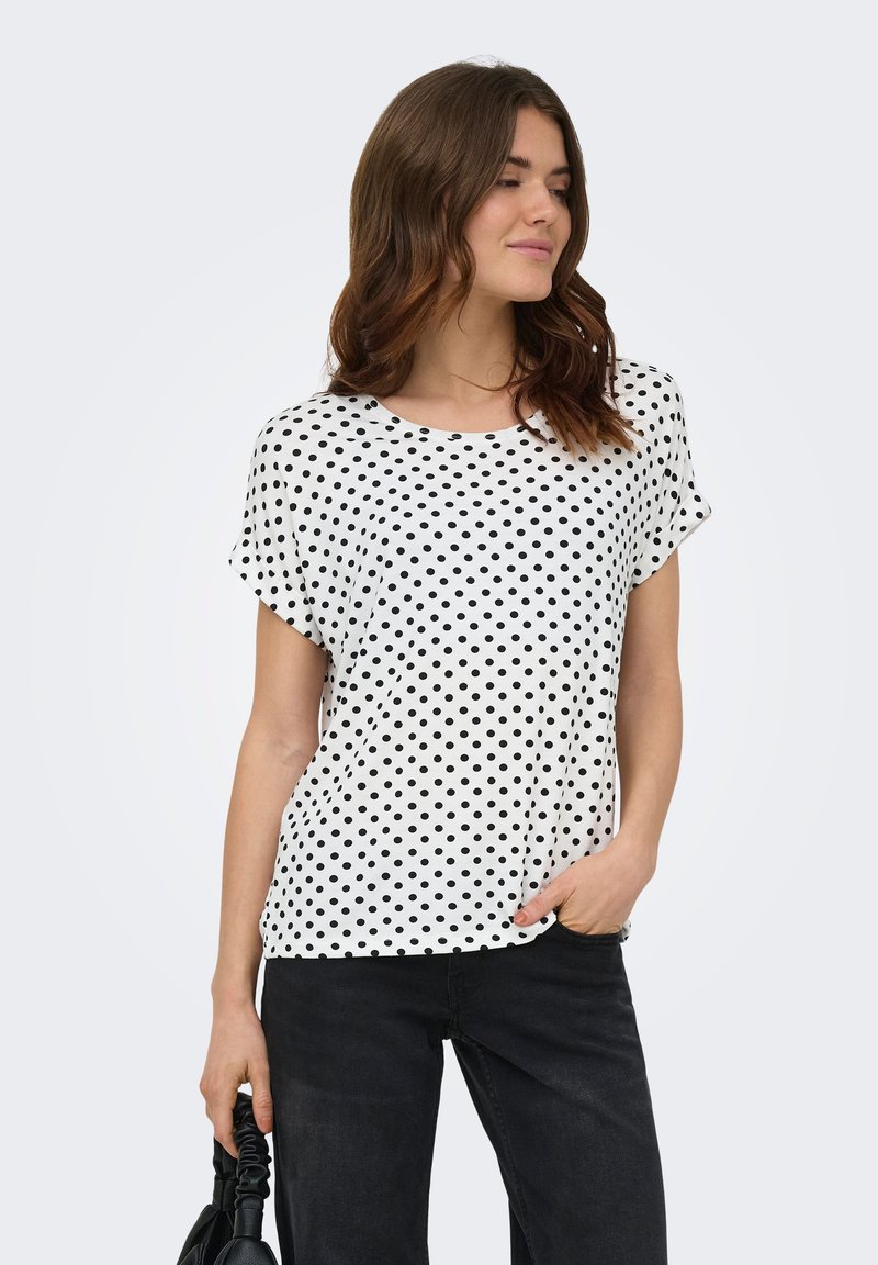 Femme aux cheveux bruns portant un chemisier blanc à manches courtes avec des pois noirs et un pantalon noir, tenant un sac à main noir dans sa main gauche.
