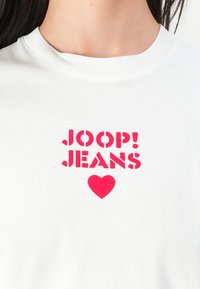 White cotton t-shirt featuring bold pink text: "JOOP! JEANS" and a heart symbol, crew neckline, smooth texture. Casual design.