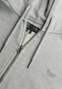 Sudadera con cremallera de color gris claro, hecha de material suave, con una cremallera delantera con herrajes de metal y el logo bordado de "Superdry" en el pecho.