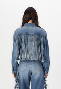 Denim jack met een franjesontwerp aan de achterkant, lichtblauwe kleur, lange mouwen en twee zakken. Heeft blauwe en witte gestreepte accenten.