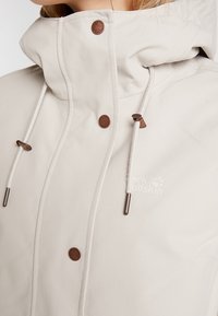 Gros plan sur une veste beige avec un col montant, des boutons pression marron, des cordons blancs et un logo discret visible sur la poitrine.