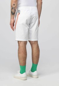 Shorts bianchi in cotone con orlo piatto, dotati di una tasca posteriore e un dettaglio in corda arancione. Indossati con calzini verdi e sneakers bianche.