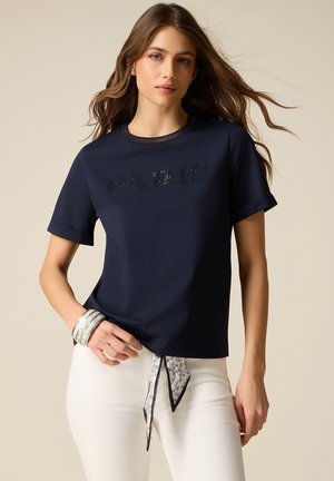 Mladá žena s dlouhými hnědými vlasy, oblečená v tmavě modrém tričku s nápisem "PARIS", bílých kalhotách s páskem z barevné šátku a stříbrnými náramky.