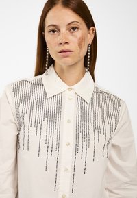 Weißes Hemd mit Knöpfen, das schwarze Strasssteine hat, die in vertikalen Linien über die Vorderseite angeordnet sind. Klassischer Kragen und perlenähnliche Knöpfe.