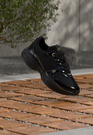 Baskets noires Tommy Hilfiger avec détails en mesh et vernis, en l'air au-dessus d'un sol en dalles de bois, cadre extérieur avec verdure et mur en pierre en arrière-plan.