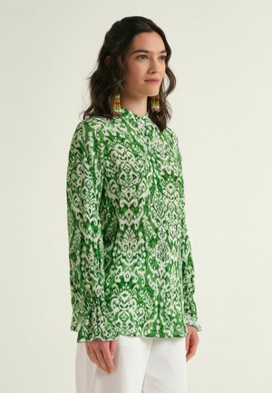 Woman wearing a green and white patterned long-sleeve blouse with ruffled cuffs and white pants, standing against a neutral background.