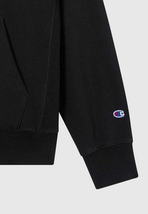Manica di felpa nera con polsino a costine e piccolo logo Champion ricamato in blu, rosso e bianco vicino al polso.