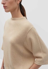 Frau mit braunen Haaren zu einem Pferdeschwanz gebunden, trägt einen beigen, kurzärmeligen Strickpullover mit hohem Rundhalsausschnitt vor einem einfarbigen Hintergrund.