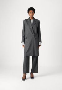 Blazer grigio a righe con rever a punta, abbinato a pantaloni coordinati e a un maglione grigio chiaro sotto. Scarpe con tacco nere.