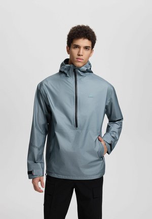 Giovane uomo che indossa una giacca a vento con cappuccio azzurro chiaro con mezza cerniera e pantaloni cargo neri, in piedi davanti a uno sfondo chiaro uniforme.