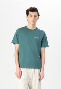 Camiseta de algodón color verde azulado con mangas cortas, cuello redondo, y texto frontal "Hotel de Palermo" en blanco. Se lleva con pantalones de color claro.