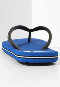 Tongs bleues avec une semelle texturée, des bretelles en caoutchouc noir en forme de cœur, et des rayures blanches et noires le long de la semelle extérieure.