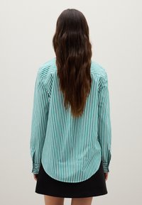 Camicia a maniche lunghe a strisce verticali turchesi e bianche, con orlo arrotondato e polsini con bottoni. Gonna nera visibile sul fondo.