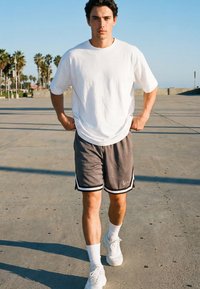 Junger Mann in weißem T-Shirt, grauen Shorts und weißen Turnschuhen, der auf einem Betonweg mit Palmen im Hintergrund unter klarem Himmel geht.