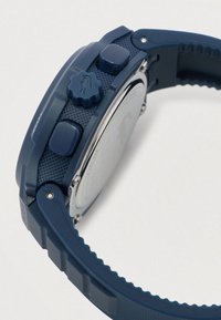 Montre numérique bleue au poignet avec des boutons texturés, un bracelet réglable strié et un dos en métal argenté, présentée en angle latéral.