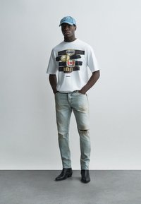 Άνδρας με ανοιχτό μπλε καπέλο, λευκό μπλουζάκι με σχέδιο, σκισμένο τζιν ανοιχτού χρώματος και μαύρες μπότες, στέκεται με τα χέρια στις τσέπες.