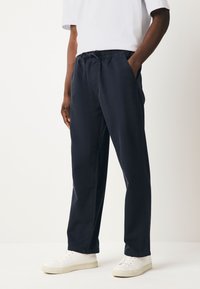 Tamsiai mėlynos medvilnės mišinys kelnės su elastinga juosmens juostele, šoniniais kišenėmis ir laisvu kirpimu, derinamos su baltais sportbačiais.