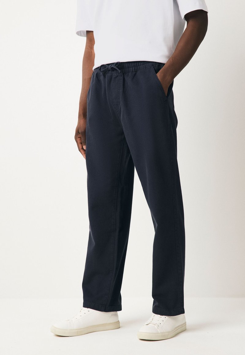 Tamsiai mėlynos medvilnės mišinys kelnės su elastinga juosmens juostele, šoniniais kišenėmis ir laisvu kirpimu, derinamos su baltais sportbačiais.