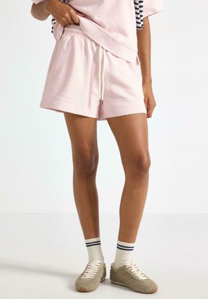 Persona che indossa pantaloncini rosa chiaro con coulisse, top abbinato, calzini bianchi con strisce scure e sneakers beige con lacci su sfondo semplice.