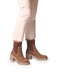 Stivaletti alla caviglia in suede marrone con polsini in maglia elastica, punta tonda e tacchi grossi; cuciture beige a contrasto e suola testurizzata.