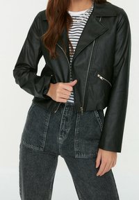 Zwarte neplederen cropped jas met een ritssluiting aan de voorkant, een notch-kraag en twee zijzakken, gedragen over een zwart-wit gestreept shirt.