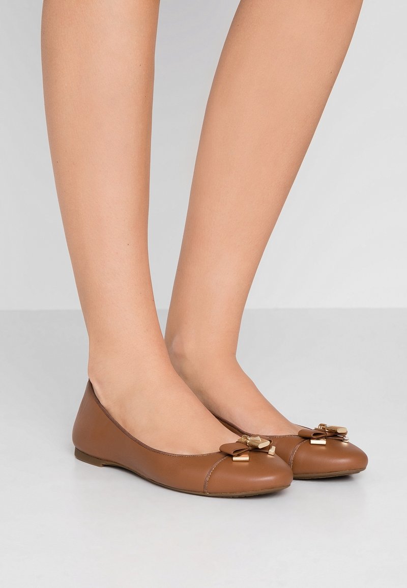 Jambes portant des ballerines en cuir marron avec des nœuds décoratifs et des ornements métalliques dorés, posées sur un sol gris clair devant un fond uni.