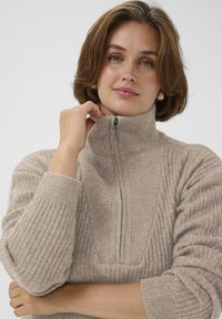 Donna con capelli corti castani che indossa orecchini a cerchio dorati e un maglione a costine beige con colletto alto zip.