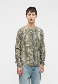 Καμουφλάζ μπλούζα με μακρύ μανίκι σε αποχρώσεις του πράσινου και του καφέ, με υφασμάτινη υφή και στρογγυλή λαιμόκοψη. Χαλαρή εφαρμογή που τονίζεται από ριχτές μανσέτες.