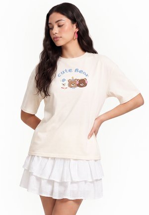 Młoda kobieta z długimi ciemnymi włosami nosi kremowy T-shirt z wizerunkami niedźwiedzi i tekstem "Cute Bear", zestawiony z białą warstwową spódnicą oraz różowymi kolczykami kołami.