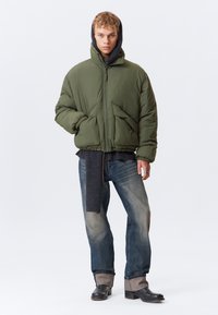 Olivgrüne gesteppte Jacke mit Frontreißverschluss, großen Taschen und Kapuze, getragen über einem dunklen Pullover und kombiniert mit blauen Jeans und schwarzen Stiefeln.